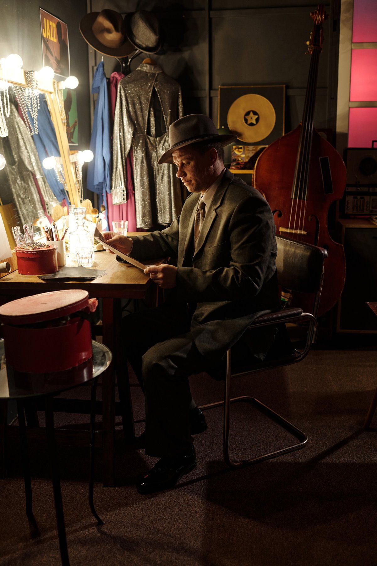 Middle aged Caucasian man wearing suit and hat sitting at dressing table reading sheet music in jazz club backstage area